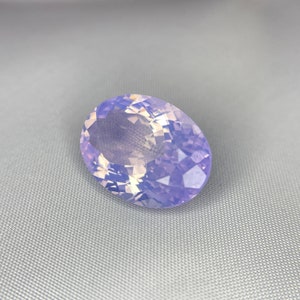 May include: A large, oval-shaped gemstone with a faceted surface. The gem has a soft, lavender color with hints of white and yellow, creating a shimmering effect. The stone is set against a gray background.