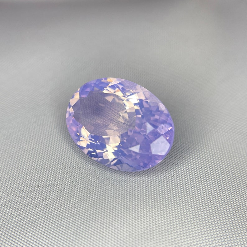 May include: A large, oval-shaped gemstone with a faceted surface. The gem has a soft, lavender color with hints of white and yellow, creating a shimmering effect. The stone is set against a gray background.