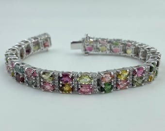 May include: A silver bracelet featuring a series of colorful, oval-cut gemstones. The gemstones are a mix of pink, green, yellow, and other hues, set in a row. Small, clear stones are interspersed between the larger gems.