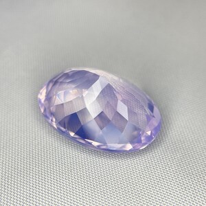 May include: A faceted, oval-shaped gemstone in a soft lavender hue. The gem's surface is cut with numerous small facets, creating a sparkling effect. The stone is set against a light gray background.