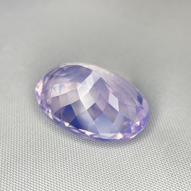 May include: A faceted, oval-shaped gemstone in a soft lavender hue. The gem's surface is cut with numerous small facets, creating a sparkling effect. The stone is set against a light gray background.