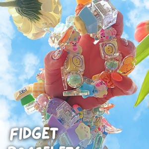 Puede incluir: Una colección de pulseras fidget coloridas con varias formas y diseños, incluyendo cubos, estrellas y personajes de dibujos animados. Las pulseras se muestran sobre un fondo de cielo azul con el texto "FIDGET BRACELETS" en blanco.