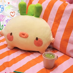 K&ouml;nnte beinhalten: Ein cremefarbenes, hasenf&ouml;rmiges Kissen mit gr&uuml;nen Ohren, rosa Wangen und einer rosa Nase. Das Kissen liegt auf einem Bett mit orange-rosa gestreifter Bettw&auml;sche. Eine Tasse gr&uuml;ne Fl&uuml;ssigkeit und bunte Marker sind ebenfalls zu sehen.