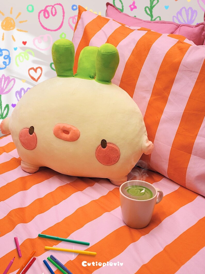 K&ouml;nnte beinhalten: Ein cremefarbenes, hasenf&ouml;rmiges Kissen mit gr&uuml;nen Ohren, rosa Wangen und einer rosa Nase. Das Kissen liegt auf einem Bett mit orange-rosa gestreifter Bettw&auml;sche. Eine Tasse gr&uuml;ne Fl&uuml;ssigkeit und bunte Marker sind ebenfalls zu sehen.