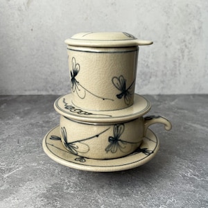 May include: Cream-colored ceramic Vietnamese coffee set with a cup, saucer, and filter. The set features a crackle glaze and is decorated with blue dragonfly motifs. The cup has a handle, and the filter has a lid.