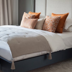 May include: A bed with a white duvet and a gray throw blanket with tassels. The bed has a headboard with several pillows in shades of orange and beige.