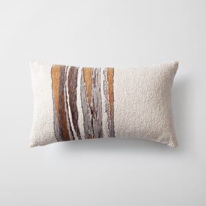 May include: A rectangular cushion with a textured, off-white fabric. Vertical stripes in shades of brown, grey, and gold decorate the centre. The cushion is designed for home decor and adds a touch of warmth and texture.