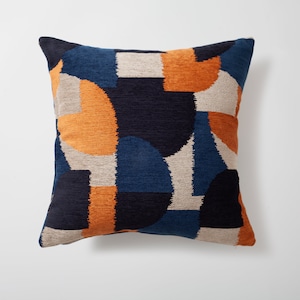 Puede incluir: Una almohada cuadrada con un patrón geométrico en tonos azules, naranjas y beige. La almohada tiene una tela suave y texturizada.