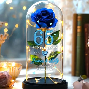 以下が含まれることがあります： ガラスドームに入った青いバラ。フェアリーライトで照らされ、「65th Anniversary」、「James & Lily」の名前、日付「09.06.1960」が記されています。黒い台座で、背景には柔らかな光と本があります。