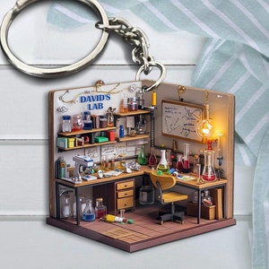 Puede incluir: Un modelo de madera en miniatura de un laboratorio de ciencias con un escritorio, estantes, vasos de precipitados y un microscopio. El laboratorio está etiquetado como "David's Lab" y tiene una luz encendida. El modelo está unido a un llavero plateado.