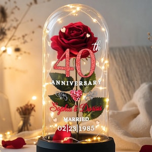 May include: A decorative glass dome with a red rose and string lights. The rose is surrounded by a 40th anniversary design with the names "Sophia & Darius" and the date "02/23/1985". The base is black.