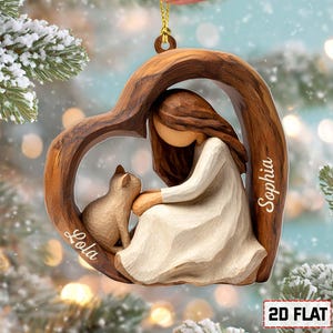 May include: A wooden heart-shaped ornament depicting a girl in a white dress, embracing a cat. The names "Lola" and "Sophia" are inscribed on the ornament. It hangs from a gold string, set against a snowy backdrop.