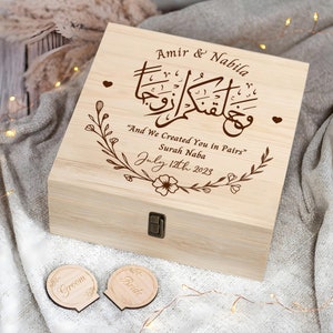 May include: A wooden box with a hinged lid, engraved with the names "Amir & Nabila" and the Arabic phrase "And We Created You in Pairs" from the Quran, Surah Naba. The box also includes the date "July 12th 2023" and a floral design. Two smaller wooden circles with the words "Groom" and "Bride" are also shown.