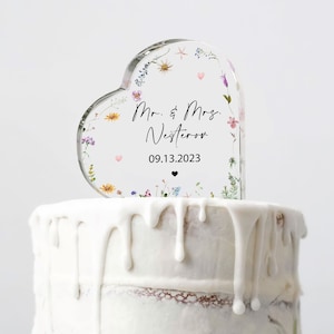 Peut inclure: Un topper de gâteau en forme de cœur transparent avec un motif floral et le texte "Mr. & Mrs. Nestorov 09.13.2023".