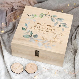 Könnte beinhalten: Eine Holzkiste mit einer hellen Holzoptik und einem Scharnierdeckel. Der Deckel zeigt ein Blumenmuster mit grünen Blättern und goldenen Akzenten. Der Text "Mr & Mrs Williams Always & Forever 30.11.2023" ist auf dem Deckel aufgedruckt. Zwei kleine Holzscheiben mit den Worten "Groom" und "Bride" befinden sich im Vordergrund.