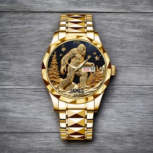 May include: A gold-toned wristwatch with a detailed Bigfoot design on the face. The watch face features a Sasquatch figure, pine trees, and stars against a dark background. The watch band is also gold-toned, and the watch reads "JAMES" at the bottom.
