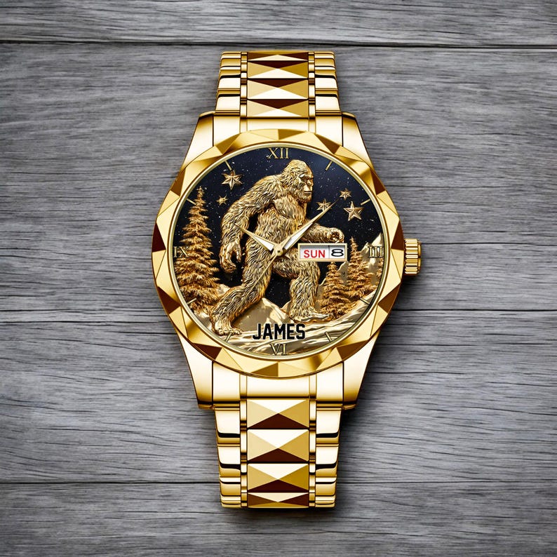 May include: A gold-toned wristwatch with a detailed Bigfoot design on the face. The watch face features a Sasquatch figure, pine trees, and stars against a dark background. The watch band is also gold-toned, and the watch reads "JAMES" at the bottom.