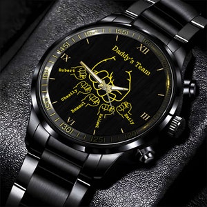 May include: Black wristwatch with a black band and a black face. The face has a gold inscription that reads "Daddy's Team" and features five fists with names written below each fist: Robert, Chasity, Besso, Kaitlynn, and Emily. The watch face also has Roman numeral hour markers.