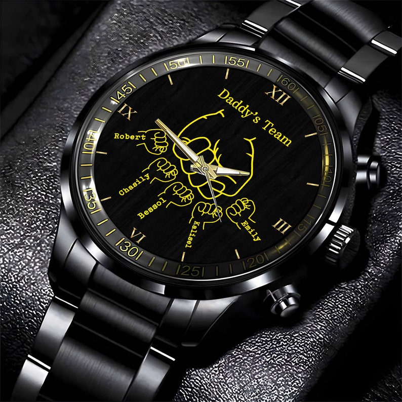 May include: Black wristwatch with a black band and a black face. The face has a gold inscription that reads "Daddy's Team" and features five fists with names written below each fist: Robert, Chasity, Besso, Kaitlynn, and Emily. The watch face also has Roman numeral hour markers.