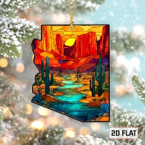 May include: Arizona-shaped ornament with a desert landscape design. The ornament features a vibrant sunset over red rock formations, cacti, and a turquoise river. The ornament is flat and has a gold string for hanging.
