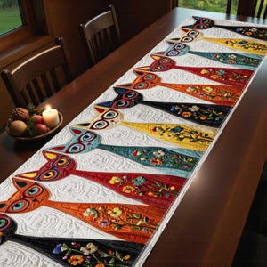 May include: A colorful, quilted table runner featuring a repeating design of stylized cat faces. The runner has a white background with various colored sections, including red, orange, yellow, and black, each adorned with floral embroidery. The cat faces have large, expressive eyes.