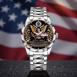 May include: A silver wristwatch with a black face featuring a golden eagle emblem, floral accents, and the text "UNITED STATES OF AMERICA." The watch also displays the date and the name "JOHNSON."
