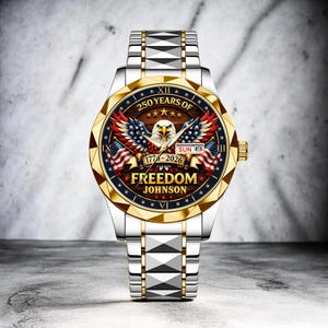 Puede incluir: Reloj de pulsera plateado y dorado con dise&ntilde;o patri&oacute;tico. La esfera del reloj presenta un &aacute;guila, banderas americanas y el texto "250 YEARS OF FREEDOM" con los a&ntilde;os 1776-2026. La correa del reloj es plateada con detalles dorados.