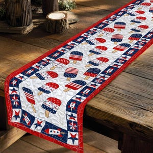 May include: A quilted table runner featuring a repeating pattern of red, white, and blue popsicle graphics on a white background. The runner is bordered by a red trim and a navy blue band decorated with white stars and small rocket ship motifs.