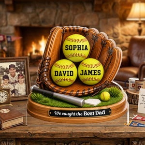 Op de afbeelding: Een gepersonaliseerde honkbaldisplay met een bruine handschoen met drie gele softballs met namen. Een honkbalknuppel, een thuisplaat en een kleine honkbal liggen op een groen veld. De basis luidt "We caught the Best Dad."