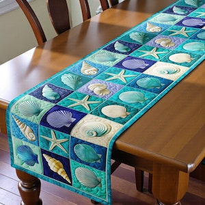May include: A rectangular table runner featuring a patchwork design with seashell and starfish motifs. The squares alternate between shades of blue, teal, and white, with a teal border. The runner is placed on a wooden table.
