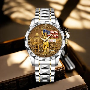 Peut inclure: Montre-bracelet argent et or avec un cadran marron repr&eacute;sentant la Statue de la Libert&eacute;, le drapeau am&eacute;ricain et le texte "UNITED STATES". Le bracelet est argent&eacute; avec des d&eacute;tails dor&eacute;s. La date est affich&eacute;e sur le cadran.