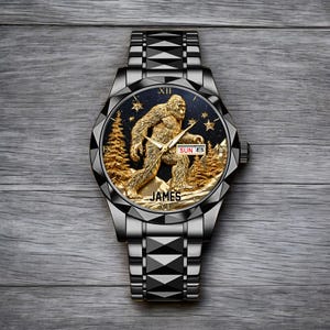 May include: A black and silver wristwatch with a gold-colored Bigfoot design on the face. The watch face features a gold Bigfoot figure, gold trees, and stars against a dark blue background. The watch band is silver and the text "JAMES" is at the bottom.