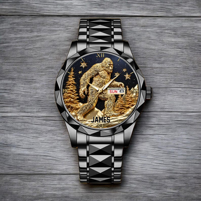 May include: A black and silver wristwatch with a gold-colored Bigfoot design on the face. The watch face features a gold Bigfoot figure, gold trees, and stars against a dark blue background. The watch band is silver and the text "JAMES" is at the bottom.