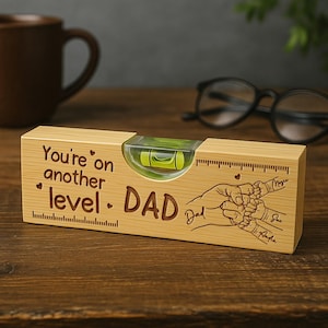 Op de afbeelding: Een houten waterpas met de tekst "You're on another level. DAD" en een afbeelding van handen die vingers vasthouden. De waterpas heeft een heldere buis met een groene vloeistofbel en centimeter markeringen.