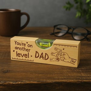 Puede incluir: Un nivel de madera con el texto "You're on another level. DAD" y un gráfico de manos y huellas de patas. El nivel tiene un vial transparente con una burbuja de líquido verde y marcas de medición.