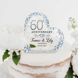Peut inclure: Un topper de gâteau en acrylique transparent en forme de cœur avec un design argenté de 60e anniversaire, des accents floraux bleus et le texte "Fames & Lily Mariés 03.06.1964".