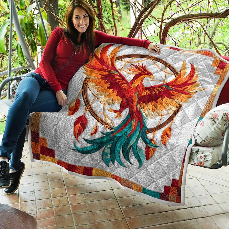 May include: A white quilt featuring a vibrant phoenix design with red, orange, and teal feathers, set against a dreamcatcher. The quilt has a geometric border in shades of red, orange, and brown. The quilt is on a swing.