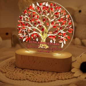 May include: A round, illuminated acrylic tree with red and white leaves, a gold trunk, and a wooden base. The text reads "40th Anniversary" with names and a date. A decorative and personalized gift.