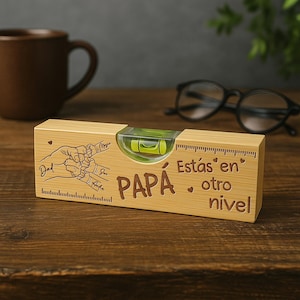 Puede incluir: Un nivel de madera con una ampolla de burbuja, grabado con la palabra "PAPA" y la frase "Estás en otro nivel." Presenta un dibujo de manos y una regla en el lateral. Una taza marrón y gafas en el fondo.