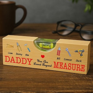 Puede incluir: Un nivel de madera con burbuja, con las palabras "DADDY MEASURE" en rojo, junto con nombres e ilustraciones de herramientas. También está presente el texto "You Are Loved Beyond". El nivel está sobre una superficie de madera.
