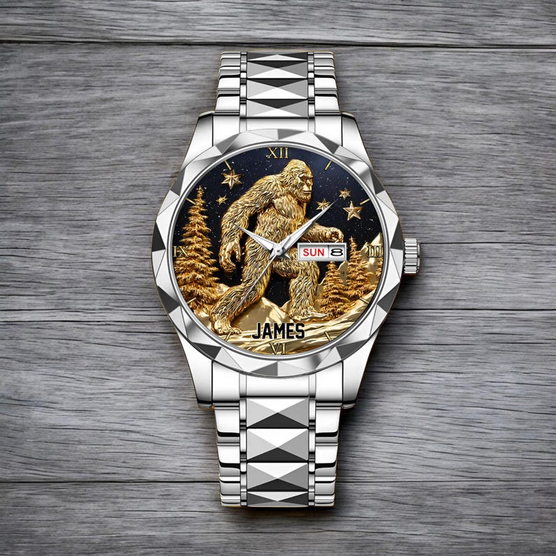 May include: Silver wristwatch with a detailed gold-colored Sasquatch design on the watch face. The watch face also features gold trees, stars, and the name "JAMES". The date is displayed as "SUN 8".
