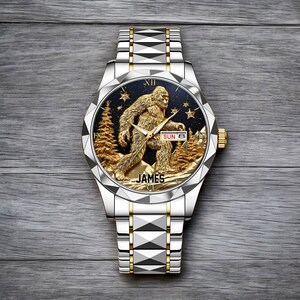 May include: Silver and gold-toned wristwatch with a detailed Sasquatch design on the face. The watch face features a gold-colored Bigfoot figure, trees, and stars against a dark blue background. The watch band is silver with gold accents, and the name "JAMES" is printed on the watch face.