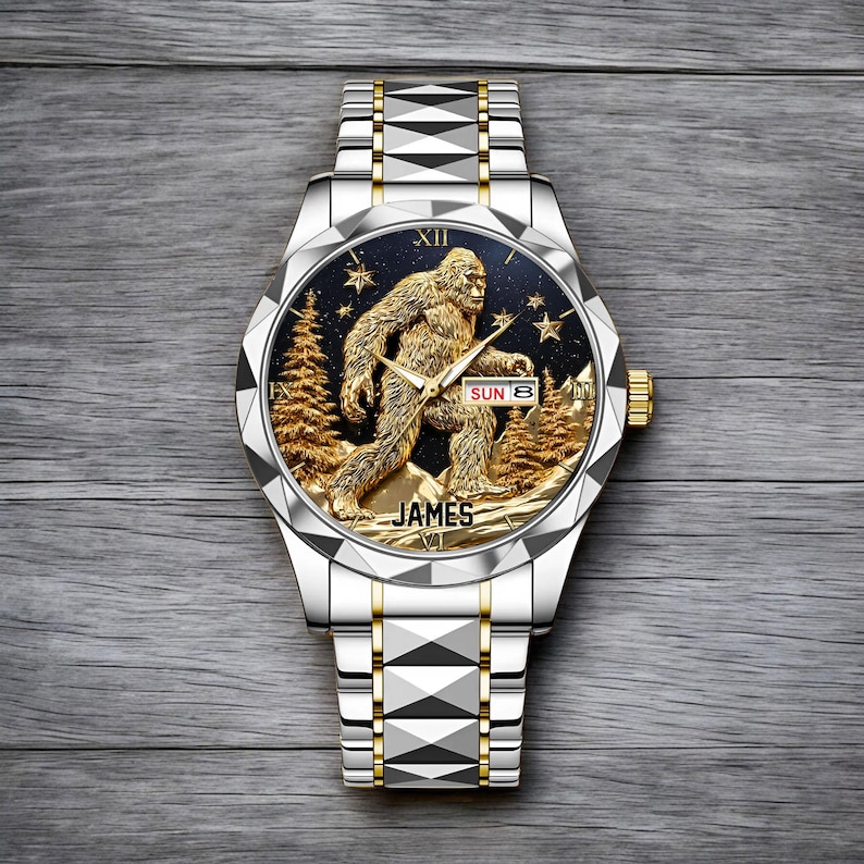 May include: Silver and gold-toned wristwatch with a detailed Sasquatch design on the face. The watch face features a gold-colored Bigfoot figure, trees, and stars against a dark blue background. The watch band is silver with gold accents, and the name "JAMES" is printed on the watch face.