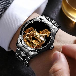May include: A silver-toned wristwatch with a black face featuring a gold-colored horse design. The watch face displays the day and date. The band is made of silver-toned metal. The watch has the name "JAMES" on the face.