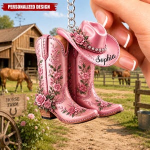 May include: A pink keychain featuring a pair of cowboy boots and a cowboy hat, all decorated with raised floral patterns and roses. The hat includes the name Sophia in black script. The design is attached to a silver metal chain link.