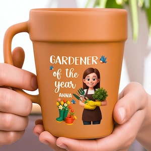 May include: A terracotta-colored ceramic mug with a handle, featuring a cartoon illustration of a gardener holding tools and a plant. The mug has the text "GARDENER of the year ANNA" and images of flowers and a watering can.