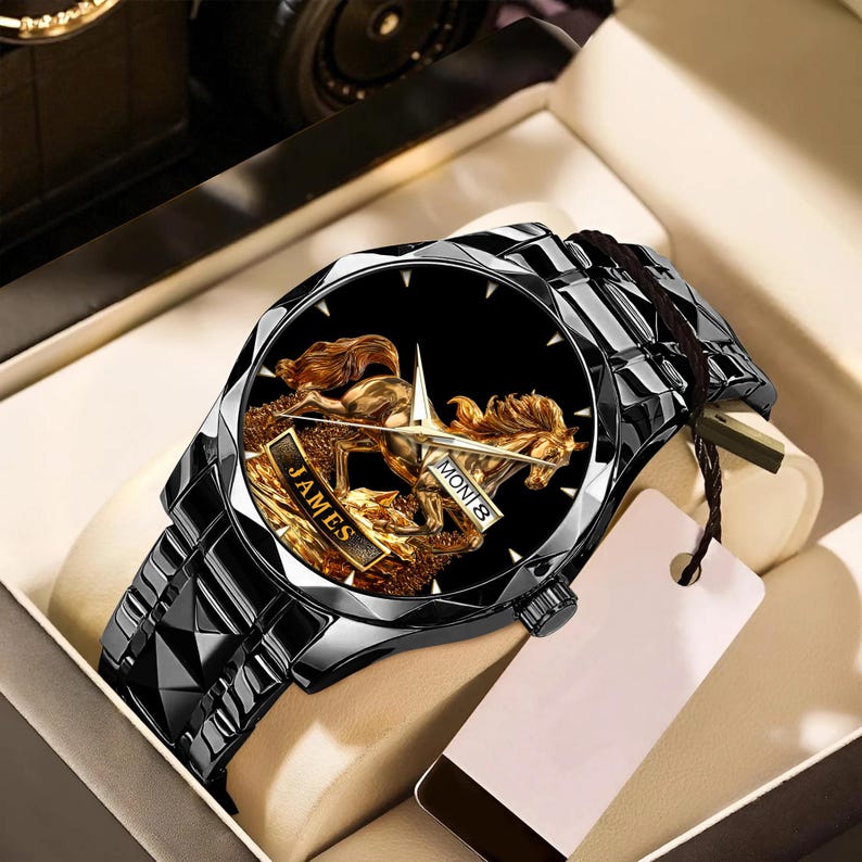 May include: Black wristwatch with a geometric design. The watch face features a gold horse and rider design with the name "JAMES" and the day of the week. The watch is presented in a box with a tag attached.