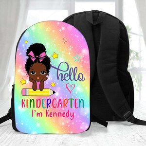 Peut inclure: Un sac &agrave; dos &agrave; motif arc-en-ciel rose, bleu et jaune avec une fermeture &eacute;clair et des bretelles noires. Le devant du sac &agrave; dos pr&eacute;sente une illustration de dessin anim&eacute; d'une fille noire avec un n&oelig;ud rose dans les cheveux assise sur un crayon jaune. Le texte "hello" et un c&oelig;ur sont au-dessus de la fille. Le texte "kindergarten I'm Kennedy" est en dessous de la fille.