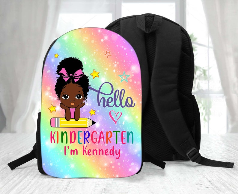 Peut inclure: Un sac &agrave; dos &agrave; motif arc-en-ciel rose, bleu et jaune avec une fermeture &eacute;clair et des bretelles noires. Le devant du sac &agrave; dos pr&eacute;sente une illustration de dessin anim&eacute; d'une fille noire avec un n&oelig;ud rose dans les cheveux assise sur un crayon jaune. Le texte "hello" et un c&oelig;ur sont au-dessus de la fille. Le texte "kindergarten I'm Kennedy" est en dessous de la fille.