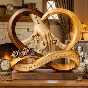 Puede incluir: Una escultura de madera con un diseño de corazón entrelazado y símbolo de infinito. La escultura incluye manos talladas que se sostienen, con los nombres "Sophia", "Alex" y "David" en los brazos. La palabra "Mommy" está inscrita en la mano más grande. La base tiene la inscripción: "To the world, you are a mum, but to us, you are our whole world."